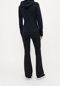 Sudadera negra con un bolsillo frontal, capucha y puños acanalados, combinada con pantalones de campana negros y zapatillas blancas. Textura de tela suave.