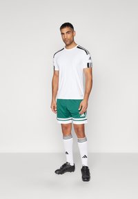 Hvit idretts T-skjorte med svarte skulderstriper, grønne shorts med hvite detaljer, svarte fotballsko og hvite sokker med svarte striper.