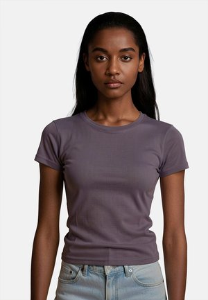Giovane donna con lunghi capelli scuri che indossa una maglietta aderente viola a maniche corte e jeans azzurri, in piedi davanti a uno sfondo semplice.