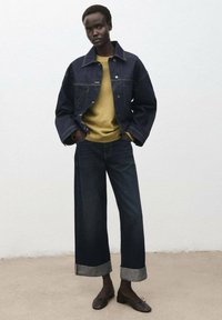 Giubbotto di jeans in tonalità scura, abbinato a un maglione giallo, jeans a gamba larga scuri con risvolti ai polsi, e ballerine marroni. Design semplice e strutturato.