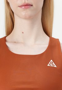 Top sin mangas de color óxido con un escote redondo y un logo triangular blanco con "acg" en el lado derecho. Textura de tela suave.