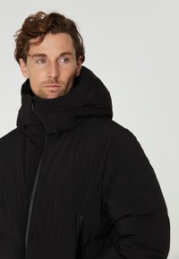Chaqueta acolchada negra con cuello alto y cierre frontal de cremallera. Presenta bolsillos laterales y una textura mate suave.