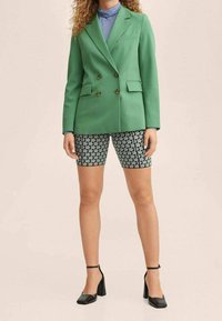 Zielona marynarka dwurzędowa, dopasowane spodenki w wzory, czarne buty na wysokim obcasie i jasnoniebieski golf, na neutralnym tle.