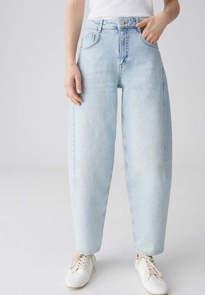 Person trägt hellblaue Jeans mit hoher Taille und weitem Bein, weiße Sneakers und ein weißes ärmelloses Top, steht vor einem schlichten Hintergrund.