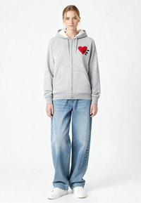 Felpa grigia con zip e motivo a cuore rosso, con dettagli neri, dotata di un morbido rivestimento, tasche frontali e cappuccio regolabile. Indossata con jeans a gamba larga blu.