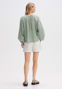 Blusa verde chiaro a maniche lunghe in tessuto testurizzato, con scollatura arricciata e vestibilità ampia, abbinata a pantaloni corti bianchi e sandali verdi.