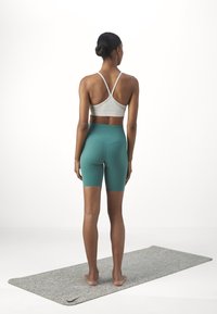 Conjunto atlético que incluye un sujetador deportivo gris claro y unos pantalones cortos de talle alto en color azul verdoso. Debajo hay una alfombra con una superficie texturizada en gris.