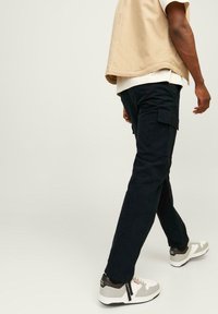 Pantaloni cargo neri con tasche laterali, abbinati a un giubbotto senza maniche beige. Le calzature presentano accenti grigi e bianchi. Tessuto di superficie liscia.