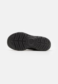 Schwarze Sneaker-Sohle mit einer strukturierten Gumm-Oberfläche, die ein Wellenmuster aufweist, und einem zentralen Logo-Emblem. Langlebig und für Rutschfestigkeit ausgelegt.