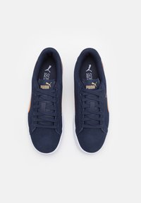 Baskets bleu marine avec tige en suede, bande latérale marron contrastante, semelle en caoutchouc blanche et lacets. Comprend une languette et une semelle intérieure portant la marque.