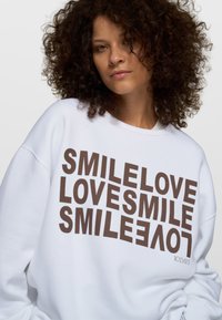 Sudadera blanca hecha de mezcla de algodón, con un texto en negrita color marrón: "SMILELOVE LOVESMILE" y un pequeño logo "10 DAYS" en el dobladillo.