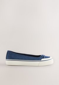 Chaussure plate slip-on en denim bleu avec semelle et embout en caoutchouc blanc, avec un petit détail nœud sur le dessus.
