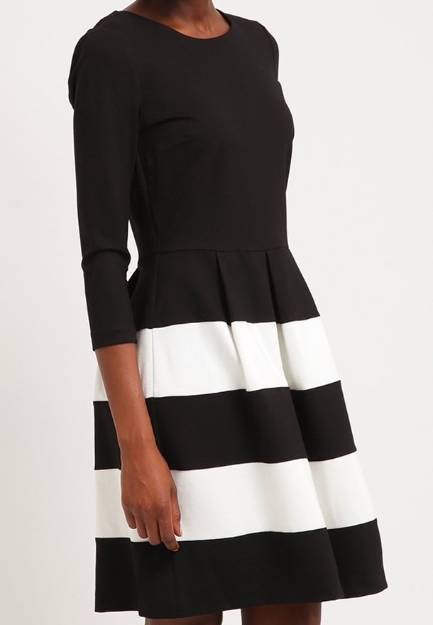 Femme portant une robe noire à manches trois-quarts et une jupe avec de larges rayures horizontales noires et blanches.