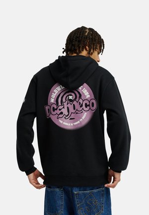 Person mit dem Rücken zur Kamera, trägt einen schwarzen Hoodie mit großem lila DC Shoes Logo und Schriftzug, kombiniert mit blauen Jeans mit Logopatches.