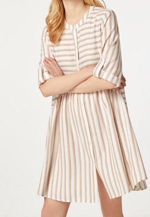 Mujer con un vestido beige y blanco a rayas verticales hasta la rodilla, con mangas remangadas y botones delanteros, brazos cruzados.