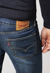 Gros plan d'une personne portant un jean Levi's 511 bleu avec un patch en cuir visible et une étiquette logo rouge sur la poche arrière.