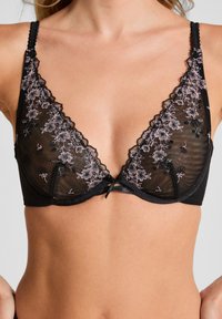 Sosten negro transparente con bordado floral en rosa y bordes en forma de olas, llevado por una mujer mostrada desde el cuello hasta el abdomen.