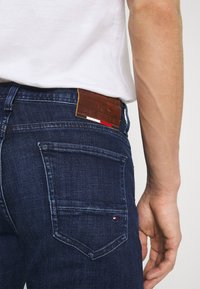 Mörkblå denimjeans med en brun läderlapp som har varumärkesnamnet och en randig accent. Femfickorsdesign med synlig sömnad.