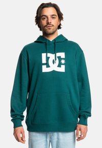 Tyrkysová mikina s kapucňou zo soft materiálu, s veľkým bielym logom DC na prednej strane, klokankovou kapsou a nastaviteľnou šnúrkou na kapucni.