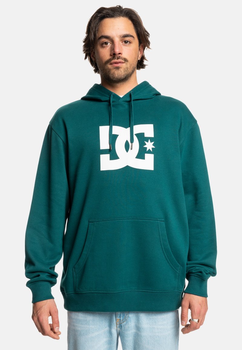 Tyrkysová mikina s kapucňou zo soft materiálu, s veľkým bielym logom DC na prednej strane, klokankovou kapsou a nastaviteľnou šnúrkou na kapucni.