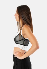 Bralette in rete nera con un design strutturato, sovrapposta a una maglietta bianca corta, indossata con una gonna nera plissettata. Tessuto liscio e leggero.