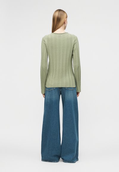 Γυναίκα με μακριά ξανθιά μαλλιά, φοράει ανοιχτό πράσινο ριμπ πουλόβερ και φαρδιά μπλε τζιν παντελόνια, στέκεται γυρισμένη πλάτη σε λευκό φόντο.