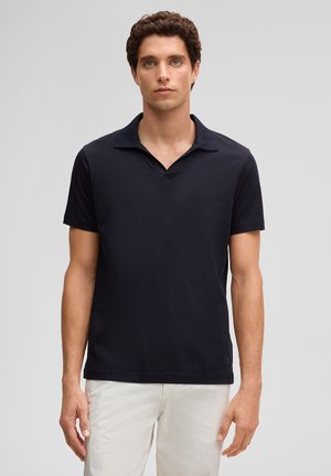 Junger Mann mit lockigem Haar, der ein schwarzes kurzärmeliges Poloshirt und weiße Hose trägt, vor einem schlichten hellgrauen Hintergrund stehend.