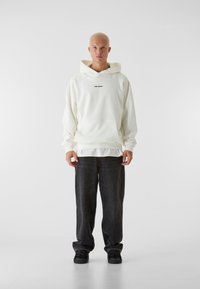 Felpa bianca con cappuccio con testo "LOST YOUR", tasca a canguro, indossata sopra una camicia bianca, abbinata a jeans neri a gamba larga e scarpe nere.