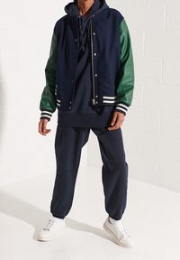 Persoon draagt een marineblauwe hoodie, een marineblauw-groene varsityjas, marineblauwe joggers en witte sneakers, staand tegen een witte muur.