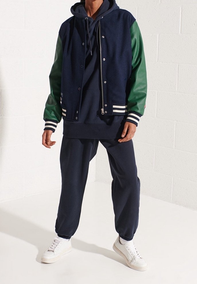 Persoon draagt een marineblauwe hoodie, een marineblauw-groene varsityjas, marineblauwe joggers en witte sneakers, staand tegen een witte muur.