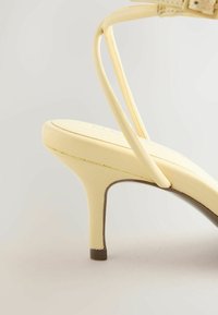 Sandale à talon haut jaune pâle avec une fine bride à la cheville et un talon aiguilles étroit sur un fond neutre.