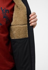 Veste noire avec une doublure en polaire beige, dotée d'une poche latérale et d'une texture lisse. La doublure intérieure est duveteuse et chaude.