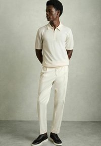 Polo bej cu mâneci scurte și guler cu fermoar, asortat cu pantaloni crema și adidași închis la culoare. Materialul pare neted, cu o croială ajustată.