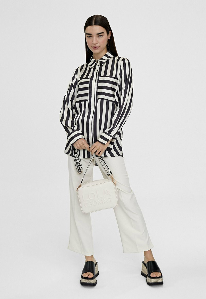 Donna che indossa una camicia a righe verticali bianche e nere, pantaloni bianchi a gamba larga, sandali neri con plataforma, mentre tiene una borsa bianca con tracolla nera.
