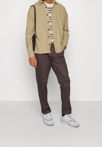 Camicia beige a bottoni sopra una t-shirt a righe, abbinata a pantaloni marroni e sneakers bianche, con una borsa a spalla nera. Design semplice.