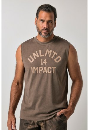 Mann mit Bart, der ein ärmelloses braunes Shirt mit dem Text "UNLMTD 14 IMPACT" trägt, steht mit einer Hand in der Tasche vor einfarbigem Hintergrund.