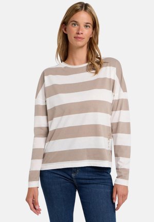 Woman wearing a beige and white wide-striped long-sleeve shirt paired with blue jeans, standing against a plain background.