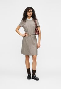 Καφέ καρό φόρεμα με κοντά μανίκια, λευκό γιακά, ζώνη στη μέση και δύο τσέπες· συνοδεύεται από μαύρα παπούτσια και καφέ κάλτσες έως τον αστράγαλο.