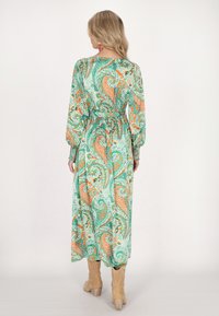 Rochie lungă cu model paisley în verde, portocaliu și crem, cu mâneci lungi, talie strânsă și fustă în straturi. Purtați cu cizme până la gleznă bej.