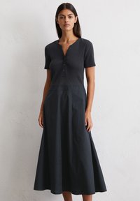A model stands against a textured white wall, wearing a dark, short-sleeved dress with a buttoned neckline and flowing skirt.