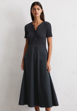 A model stands against a textured white wall, wearing a dark, short-sleeved dress with a buttoned neckline and flowing skirt.