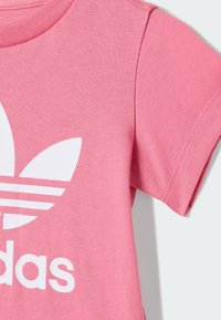 Pink cotton t-shirt with short sleeves, featuring a large white Adidas logo and three white stripes on the front, textured fabric.