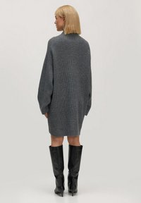 Graues, oversized Strickkleid mit hohem Ausschnitt, langen Ärmeln und lockerer Passform, kombiniert mit kniehohen schwarzen Lederstiefeln.