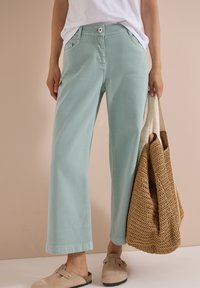 Personne portant un pantalon large vert menthe, une chemise blanche, des sabots beiges et tenant un sac cabas tressé à anse crème.