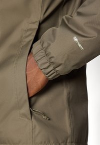 Veste imperméable vert olive avec des poignets élastiques et une poche zippée, arborant un logo "DRY VENT" sur la manche. Texture de tissu lisse.