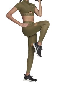 Person i olivgrön Adidas-träningskläder och svarta Adidas-sneakers som lyfter en hantel och höjer ett knä framåt i en träningsposition.