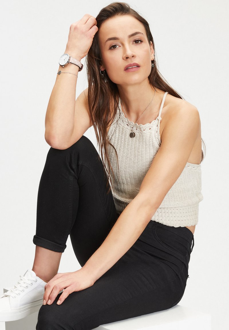 Débardeur en tricot sans manches beige avec des bords en dentelle, associé à un jean slim noir et des baskets blanches. Comprend une montre en argent et des bijoux.