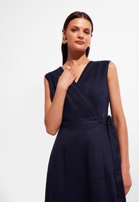 Granatowa, bezrękawnikowa sukienka o kroju kopertowym z teksturowanym wycięciem w kształcie litery V, bocznym wiązaniem i okrągłym akcentem. Gładki materiał z delikatnym połyskiem.