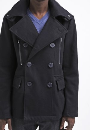 Une personne porte un manteau noir croisé à larges revers et à poches zippées, par-dessus un pull bleu à col en V.