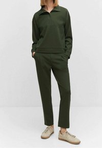 Sudadera verde oliva con cuello de cremallera hasta un cuarto, combinada con pantalones ajustados a juego. El material tiene una textura suave y lisa. Zapatillas beige completan el conjunto.